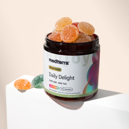 A dark amber jar labeled "Daily Delight CBD Gummies" from Medterra, filled with high potency CBD gummies, sits on a white surface with a light background. Some colorful, sugar-coated wellness gummies are scattered next to the jar.