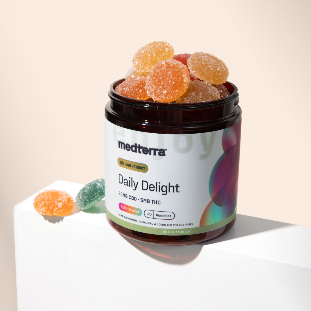 A dark amber jar labeled "Daily Delight CBD Gummies" from Medterra, filled with high potency CBD gummies, sits on a white surface with a light background. Some colorful, sugar-coated wellness gummies are scattered next to the jar.
