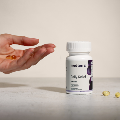 A person's hand holding a softgel capsule extends above a bottle of Medterra Daily Relief CBD Isolate Capsules, which are infused with organic MCT Coconut Oil, on a light-colored surface. The bottle is white with a purple label and additional softgel capsules are lying nearby.