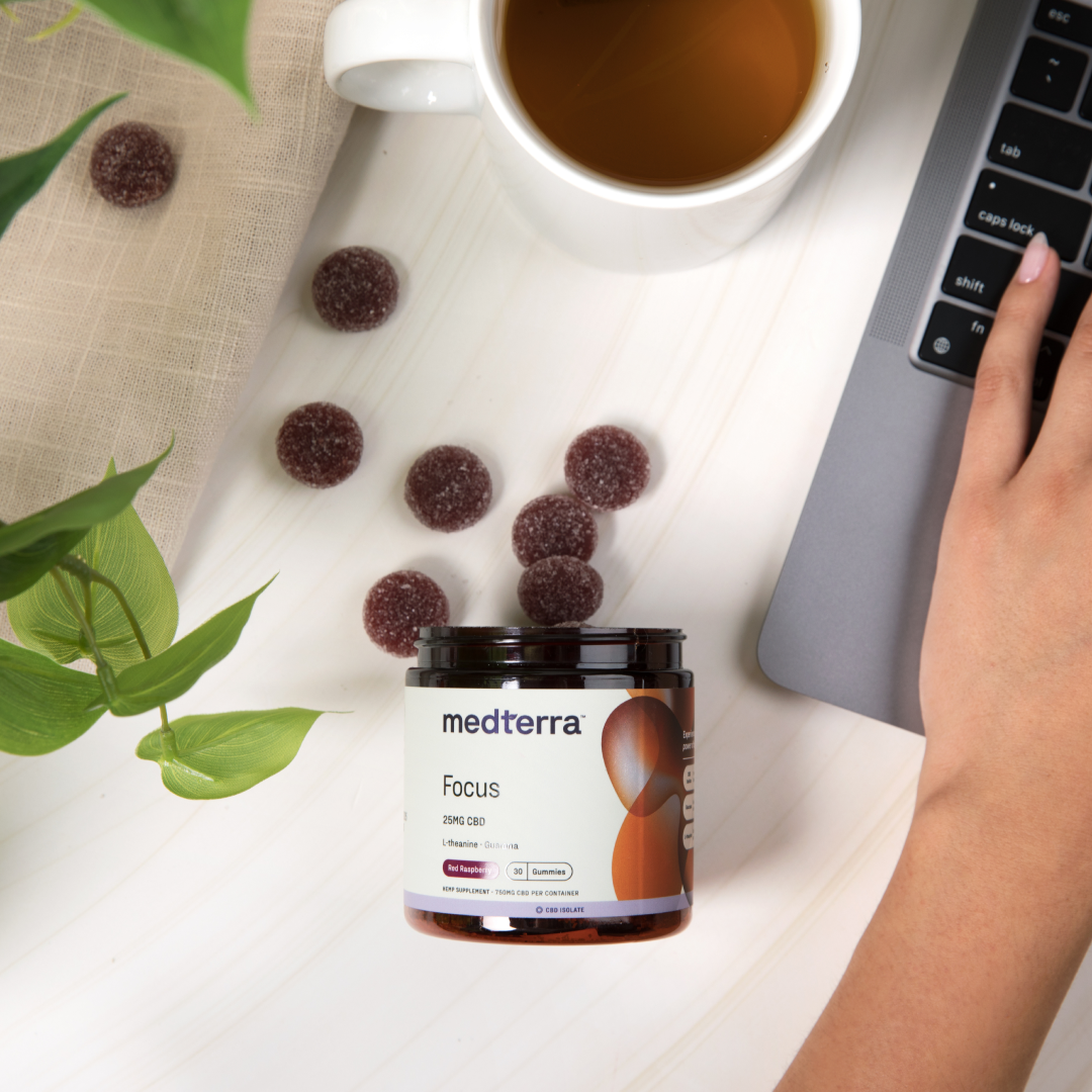 A person using a laptop with a jar of Medterra's Focus CBD Gummies beside them. Several gummies are spilled on the table. A cup of tea, a green potted plant, and a beige cloth create a cozy workspace, perfect for cognitive enhancement and sustained energy.