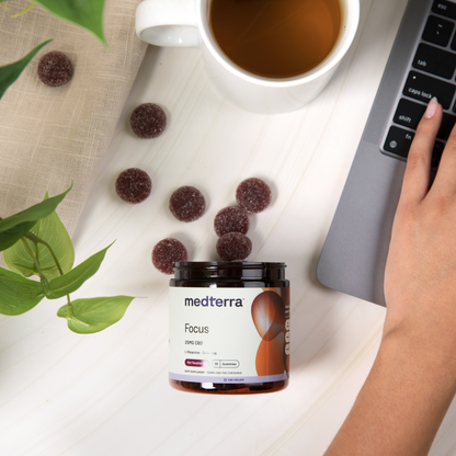 A person using a laptop with a jar of Medterra's Focus CBD Gummies beside them. Several gummies are spilled on the table. A cup of tea, a green potted plant, and a beige cloth create a cozy workspace, perfect for cognitive enhancement and sustained energy.