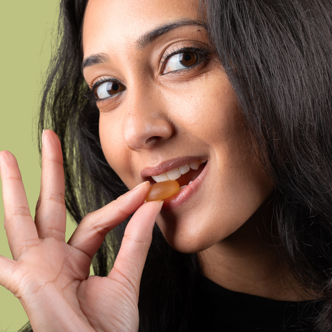 A close-up of a woman with long dark hair holding a Max Relief Full Spectrum CBD Gummy from Medterra near her lips with one hand. She is about to eat the gummy, and her other hand is making an 'OK' gesture. The background is a light green color.