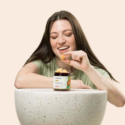 A smiling woman with long hair holds a small gummy near her mouth. In front of her is a speckled stone surface with a jar labeled "Mood Boost CBD Gummies" from Medterra. The background is a solid off-white color.