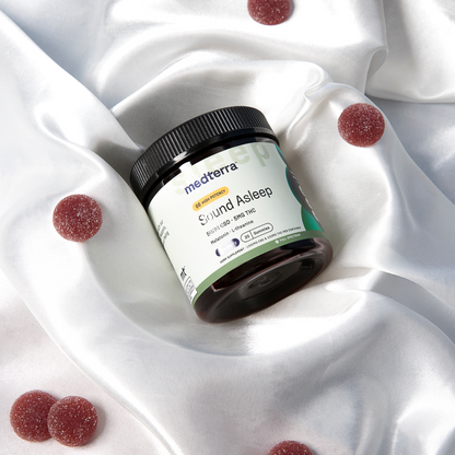 A jar labeled "Medterra Sound Asleep CBD Gummies" rests on silky white fabric surrounded by red THC gummies. The jar features a white and green label with text describing the product as a sleep aid containing CBD, CBN, and melatonin supplement for a restful night.