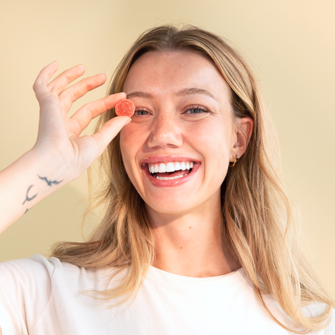 A smiling woman with long blond hair holds a small, round Daily Delight CBD gummy from Medterra up to her right eye. She has a tattoo of birds on her left forearm and is wearing a white t-shirt. The background features a muted yellow-beige color.