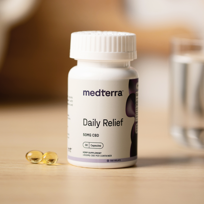 A bottle of Medterra's Daily Relief CBD Isolate Capsules, containing 30 capsules with a dosage of 50mg each, is presented in a white bottle adorned with a purple label. Three yellow CBD capsules are shown alongside it, against the backdrop of a blurred glass of water on a wooden surface, emphasizing its organic MCT Coconut Oil blend.