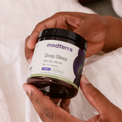 A person holds a dark amber jar labeled "Medterra Deep Sleep CBD Gummies" in their hands. The jar contains 30 gummies, each with 25mg CBD, 2mg THC, and melatonin, crafted from Full Spectrum Hemp Extract in a mixed berry flavor. The person's hands and part of a white cloth are visible in the background.