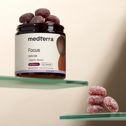 A jar of Medterra Focus CBD Gummies rests on a glass shelf. The jar is labeled "Medterra Focus, 25MG CBD, L-Theanine + Guarana, Red Raspberry, 30 Gummies." A few gummies are displayed on a nearby glass shelf, promising cognitive enhancement and sustained energy.