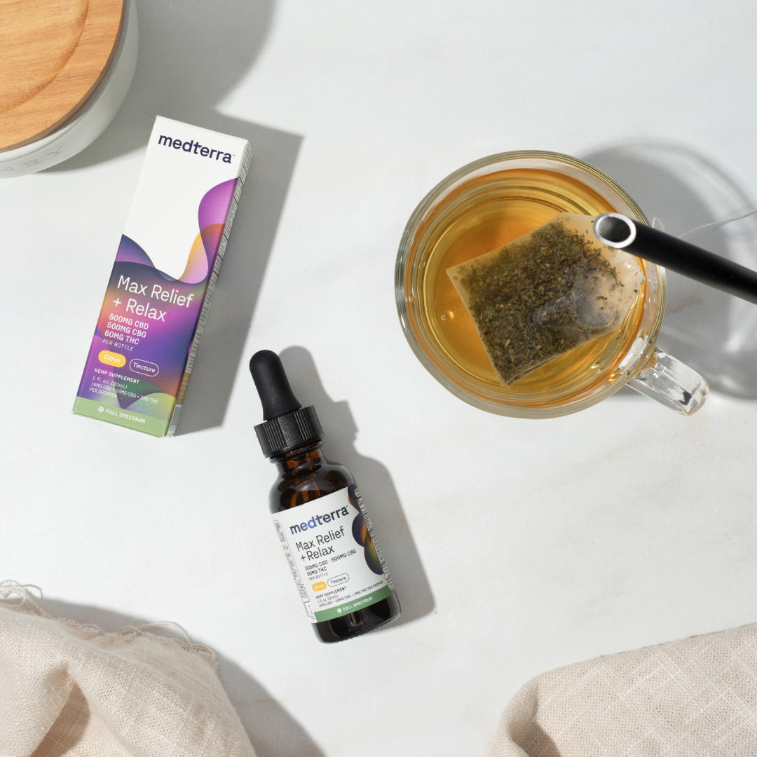 A bottle of Medterra Max Relief + Relax Full Spectrum CBD Oil with a dropper sits beside its packaging. Nearby, a glass mug with a tea bag steeping in it accompanies a beige cloth napkin, all elegantly arranged on a light-colored surface to evoke relaxation support and the entourage effect.