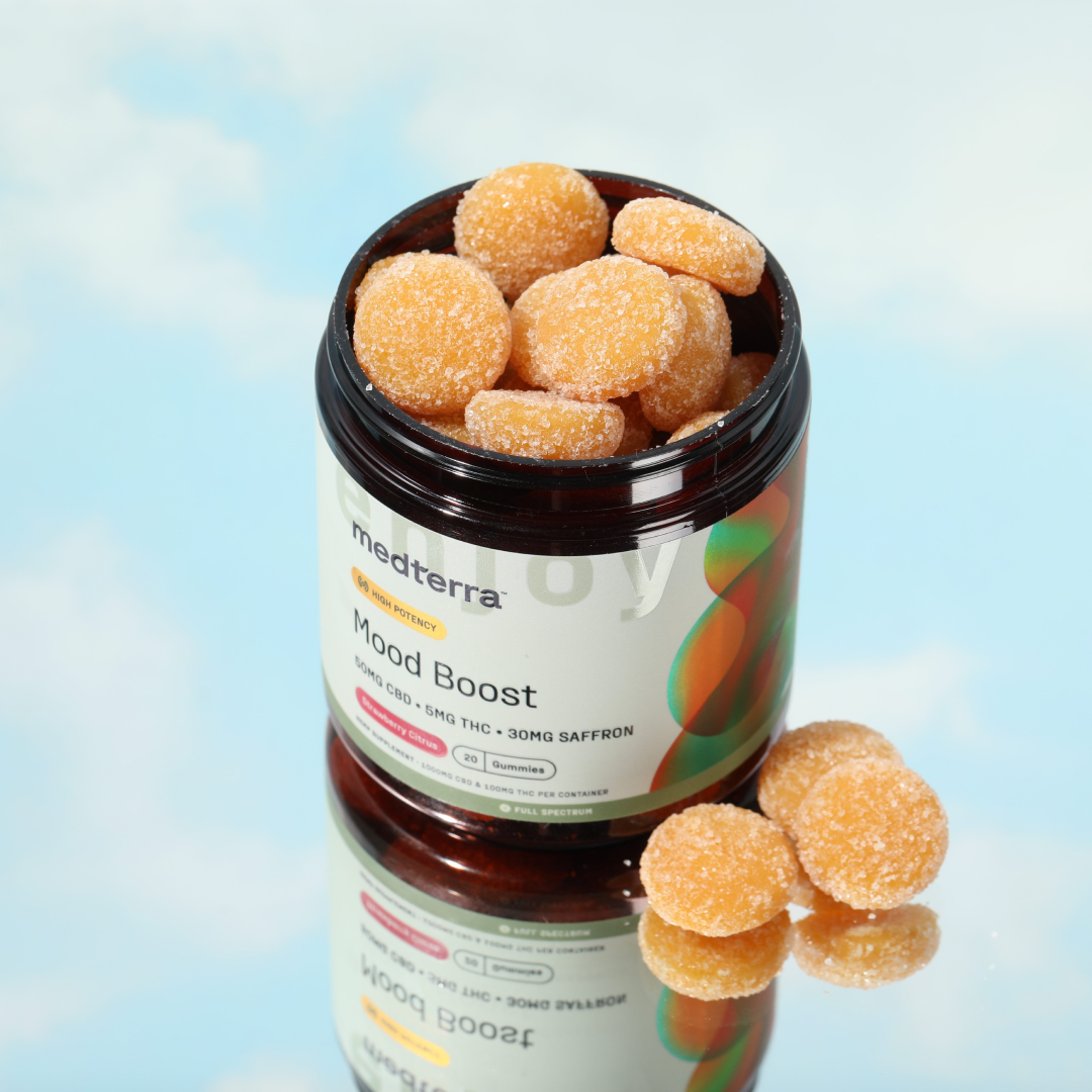 A jar of Medterra Mood Boost CBD Gummies sits open, revealing orange gummies dusted with sugar. The jar's label indicates the product contains 30 saffron-infused gummies, each with CBD and 5mg THC. It is reflected on a glossy surface with a sky-blue background.