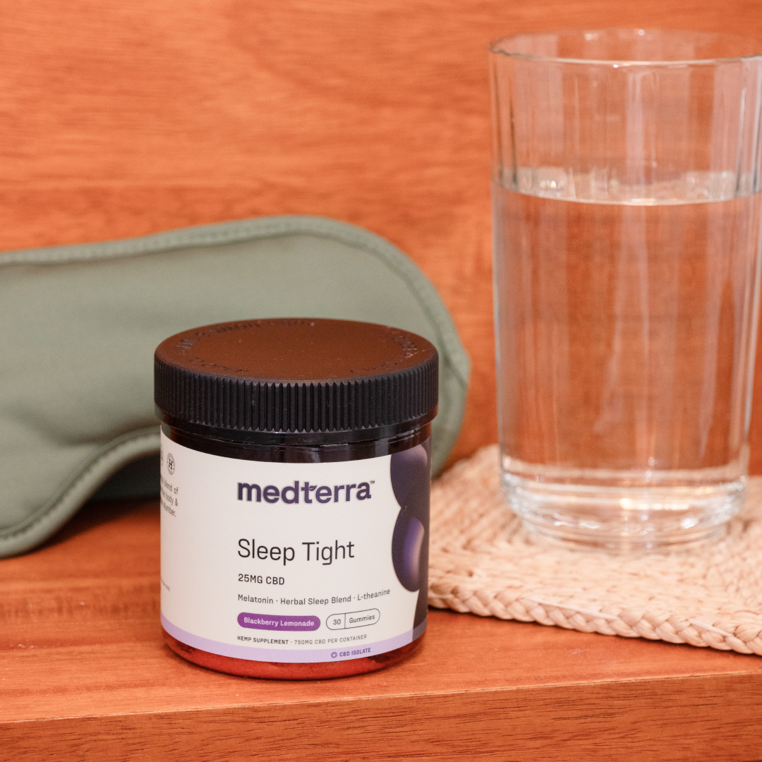 A jar of Medterra Sleep Tight CBD Gummies is placed next to a glass of water and a green sleep mask, all set on a wooden surface. The label on the jar mentions "25mg CBD" and "Blueberry Lemonade" as the flavor, promising melatonin-infused sleep support.