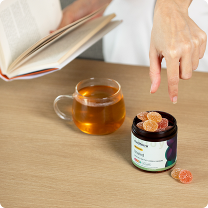 A person points to a jar of Medterra's Unwind CBD Gummies on a wooden table. Next to the jar is a clear glass mug filled with tea, and the person is holding an open book in their other hand.