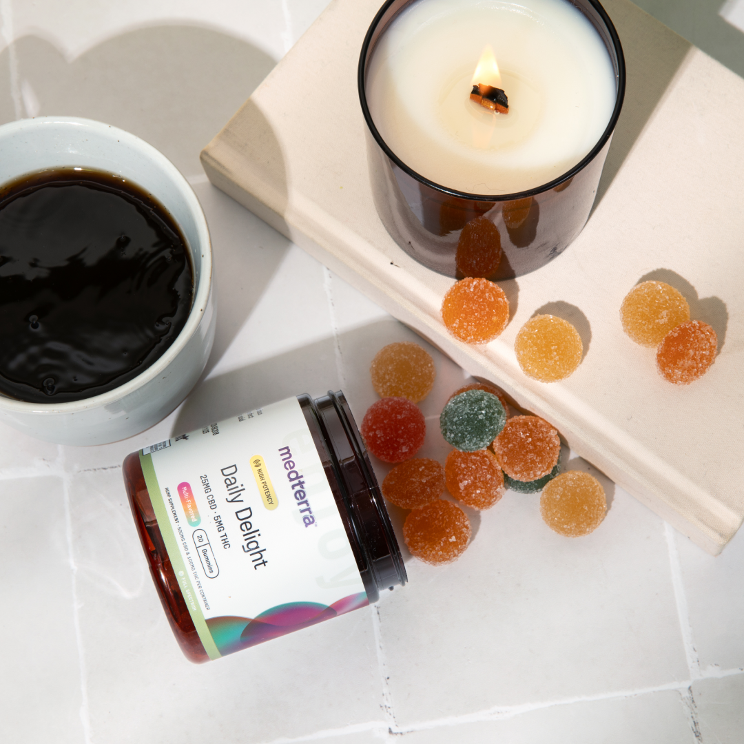 A mug of black coffee sits on a white tiled surface next to an open container of Medterra's Daily Delight CBD Gummies. The gummies are scattered beside a lit candle in a brown glass holder and a closed book, creating a cozy and inviting scene.
