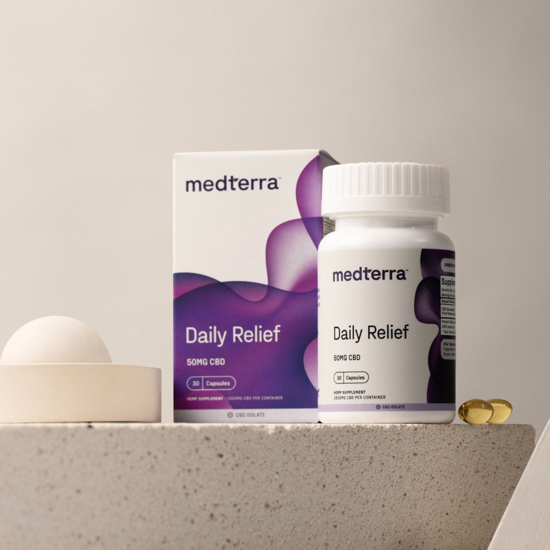 A Medterra Daily Relief CBD Isolate Capsules bottle and box are showcased on a beige background. The open bottle reveals two yellow capsules beside it. The box, adorned with a purple abstract design, prominently displays "Daily Relief 50mg CBD" and emphasizes the addition of organic MCT Coconut Oil for improved effectiveness.