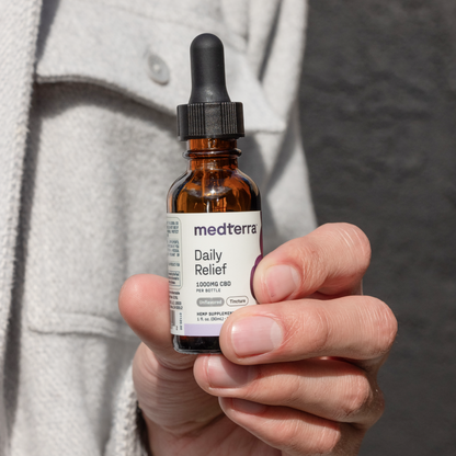 A person in a light-colored jacket holds a small brown bottle of Medterra Daily Relief CBD Isolate Oil, labeled with "1000mg Isolate CBD per bottle" and "1 fl oz / 30 ml." The bottle features a black dropper cap, and the background is a neutral, dark shade.