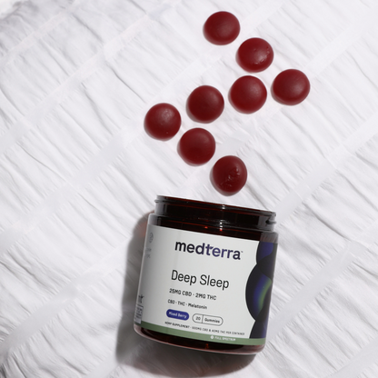 A jar of Medterra Deep Sleep CBD Gummies is partially open with several red gummies spilling out onto a white, textured surface. The product label indicates it contains 25mg CBD, 2mg THC, melatonin, and Full Spectrum Hemp Extract and is flavored as wild berry.