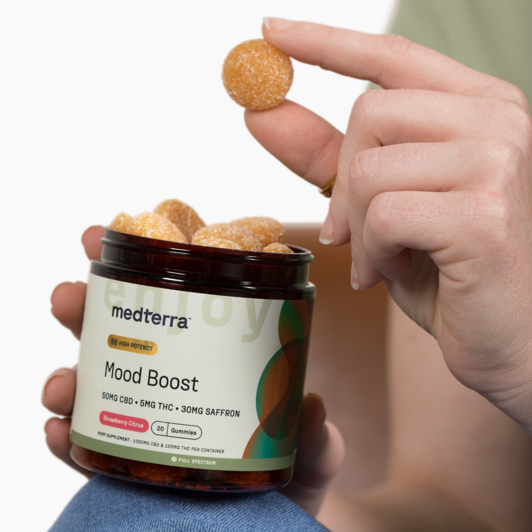 A hand holds a gummy over an open jar of Medterra Mood Boost Gummies, which are labeled with 50mg CBD, 5mg THC, and 30mg saffron. The jar indicates the flavor as Strawberry Citrus. Another hand is seen holding the jar from the side.