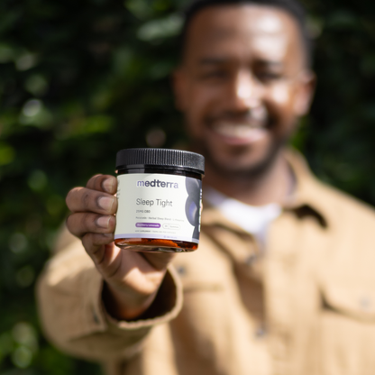A man in a brown jacket, out of focus, holds a Medterra Sleep Tight CBD Gummies jar towards the camera. The product label is clearly visible, featuring the brand and product name against a green outdoor background, highlighting its role in sleep support.