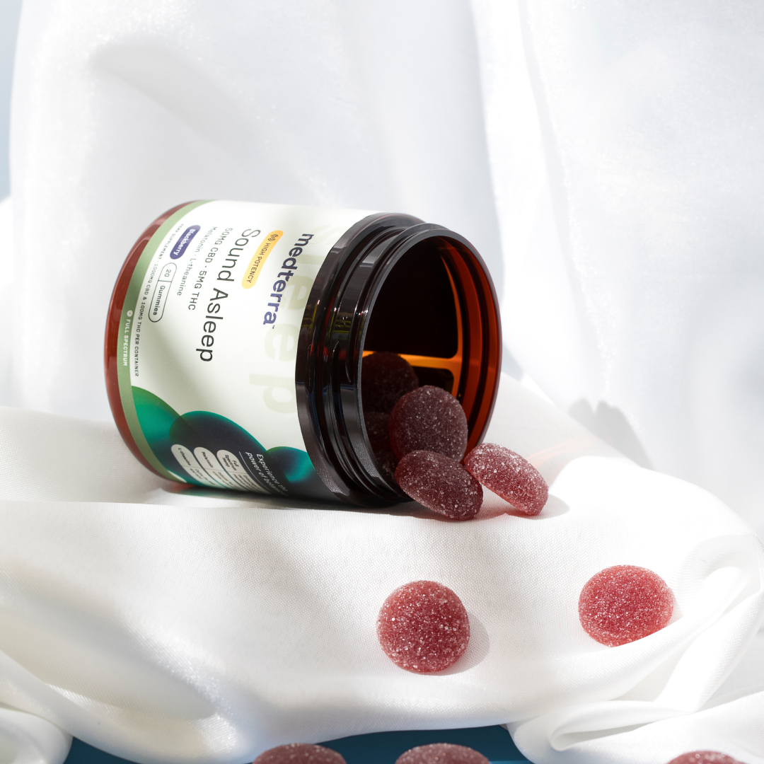 An open jar of Medterra Sound Asleep CBD Gummies with several red gummies spilling out onto a white, silky fabric background. The jar's label is prominently visible, featuring the product name and details. The scene suggests relaxation and nighttime use, ideal sleep aids for a restful night.