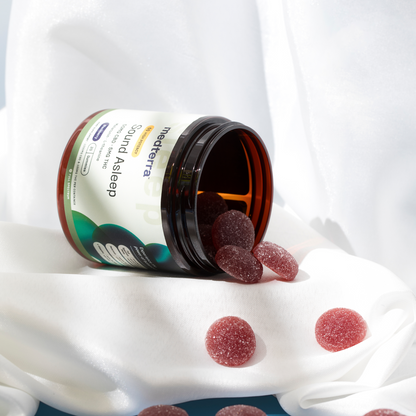 An open jar of Medterra Sound Asleep CBD Gummies with several red gummies spilling out onto a white, silky fabric background. The jar's label is prominently visible, featuring the product name and details. The scene suggests relaxation and nighttime use, ideal sleep aids for a restful night.