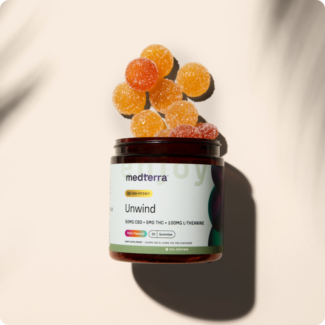 A jar labeled "Unwind CBD Gummies" by Medterra contains orange and red gummy supplements for stress relief, with ingredients listed as 50mg CBD, 5mg THC, and 100mg L-Theanine. Several sugar-coated gummies are scattered around the open jar, casting a shadow in the background.