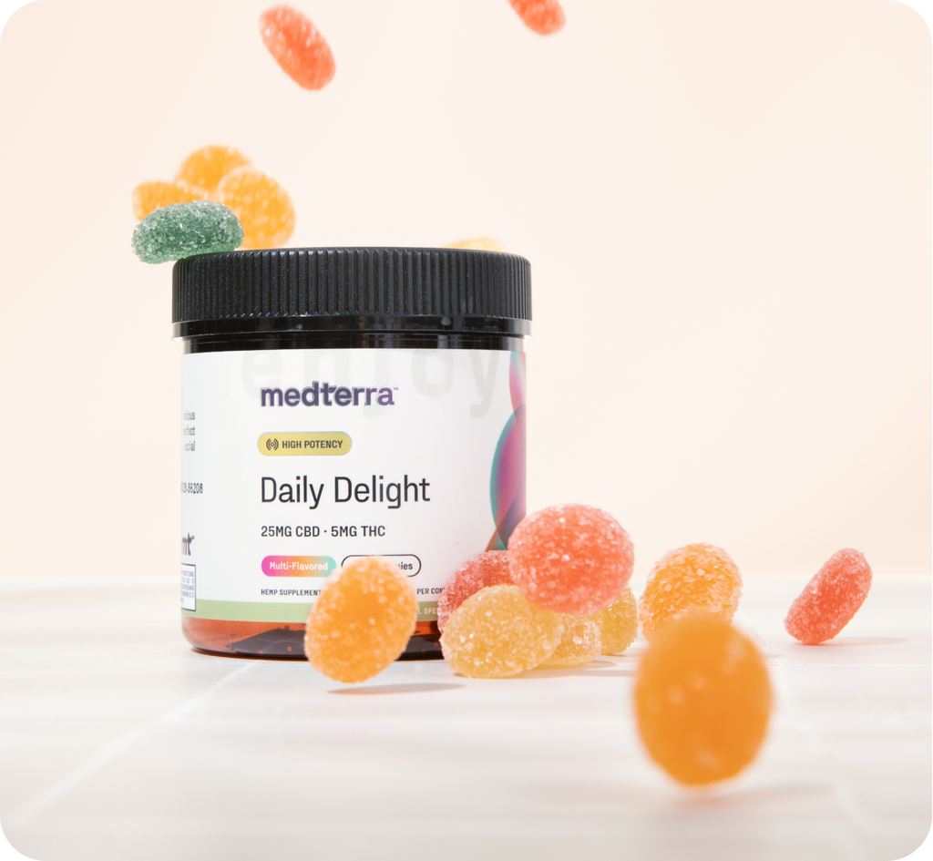A jar of Medterra Daily Delight CBD Gummies is open, with colorful gummies in various shapes and sizes scattered around it. The label indicates that each serving contains 25mg of CBD and 5mg of THC. Some gummies are also seen floating above the jar against a light background.