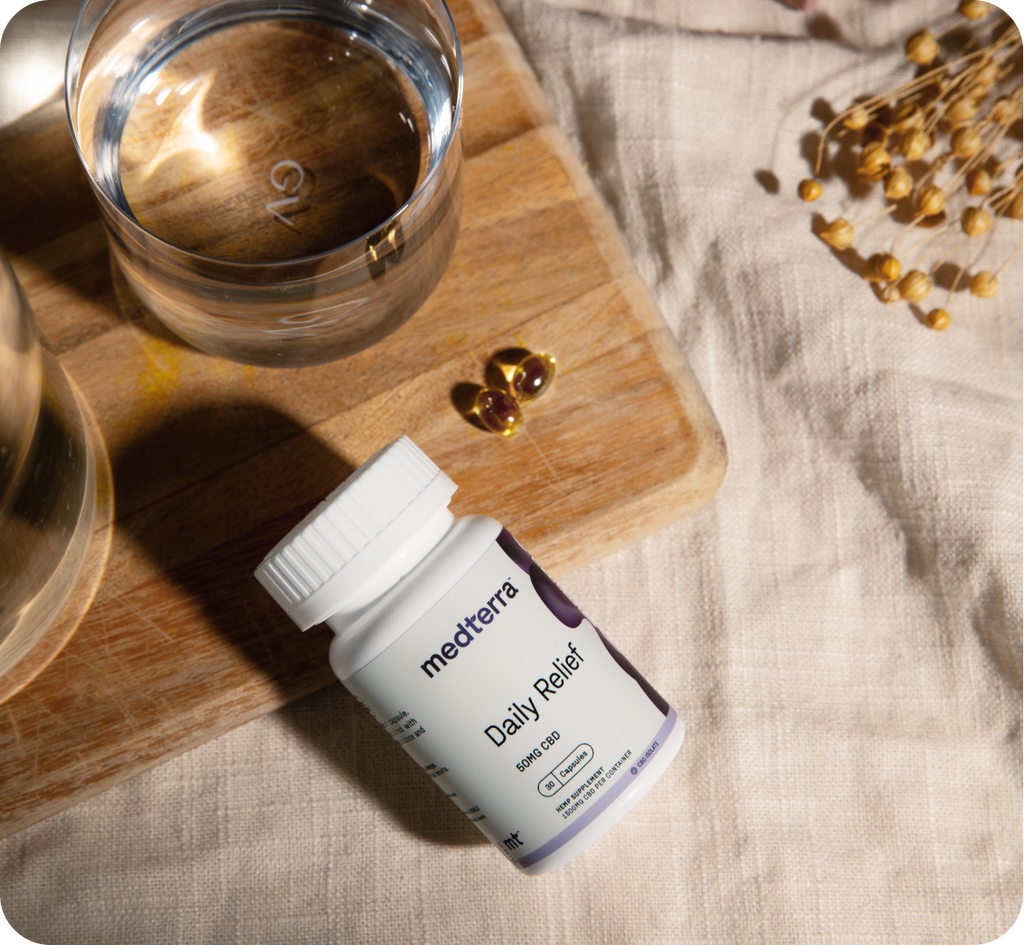 A bottle of Medterra Daily Relief zero THC CBD capsules lies on a wooden board next to a glass of water. Two capsules are placed nearby. The setup is on a light fabric surface, with a small cluster of dried flowers in the background. Perfect for on-the-go relief and shipping to all 50 states.