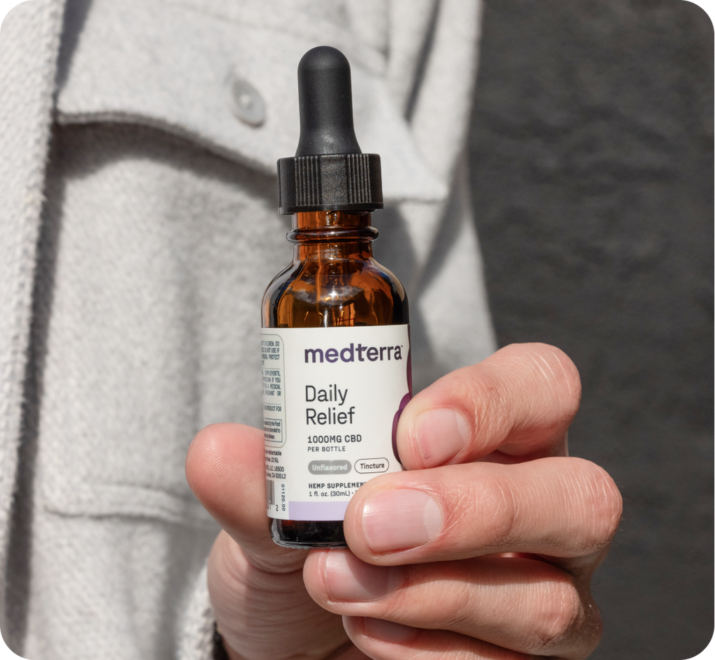 Close-up of a person holding a small brown glass dropper bottle labeled "medterra Daily Relief." The label notes "1000mg pure cannabidiol extract per bottle," "1 fl oz (30ml)," and "Tincture." The person, wearing a light-colored jacket, is showcasing the CBD oil drops.