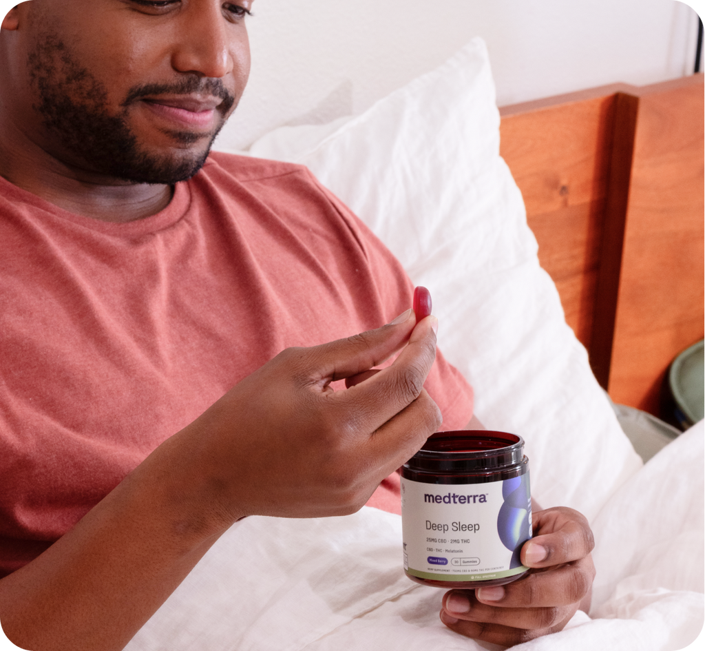 A person wearing a red shirt is sitting in bed holding a container of "Medterra Deep Sleep" supplements in one hand and a single melatonin supplement in the other, looking at it closely. The bed has white bedding, and a wooden headboard is partially visible in the background.