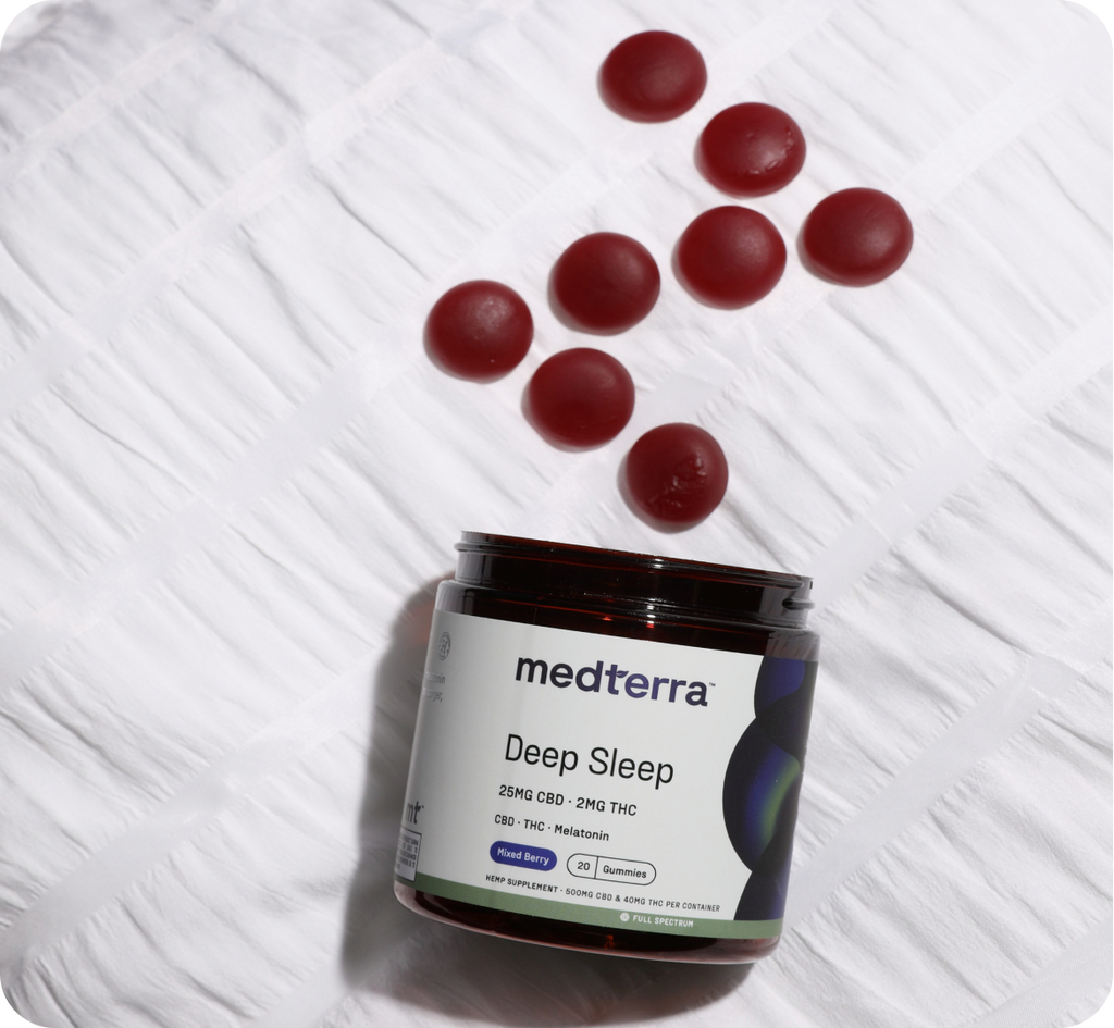 A jar labeled "medterra Deep Sleep Gummies" containing Full Spectrum Hemp Extract and melatonin is placed on a white quilted surface. Seven red gummies are scattered next to the jar, each offering 25mg CBD, 2mg THC, and a delightful "Mixed Berry" flavor.