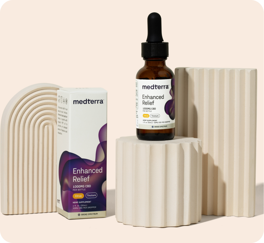 A bottle of Medterra Enhanced Relief CBD oil drops sits on a beige, ribbed pedestal. Next to it is the product box, designed with purple and teal abstract patterns. Both are surrounded by stylish, beige geometric structures on a neutral background, promising balance and calm in 1000mg and 2000mg options.