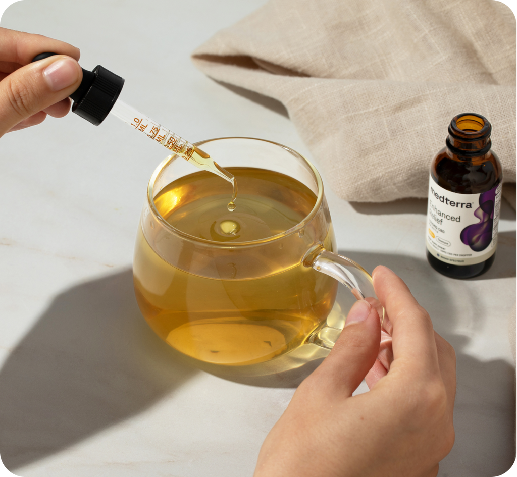 A person adds CBD oil drops from a dropper into a glass cup of tea. Nearby, an open brown bottle labeled with "1000mg and 2000mg" and another dropper cap rest on a white surface. A beige cloth serves as the background, creating a scene of balance and calm.