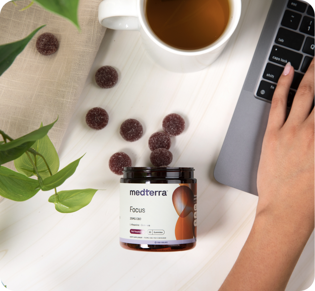 An open container of Medterra Focus gummies sits on the table, with several gummies spilled out. A cup of tea and a partially visible laptop are also present. A hand types on the laptop, clearly working to enhance their attention span, while a green plant adds a touch of nature nearby.