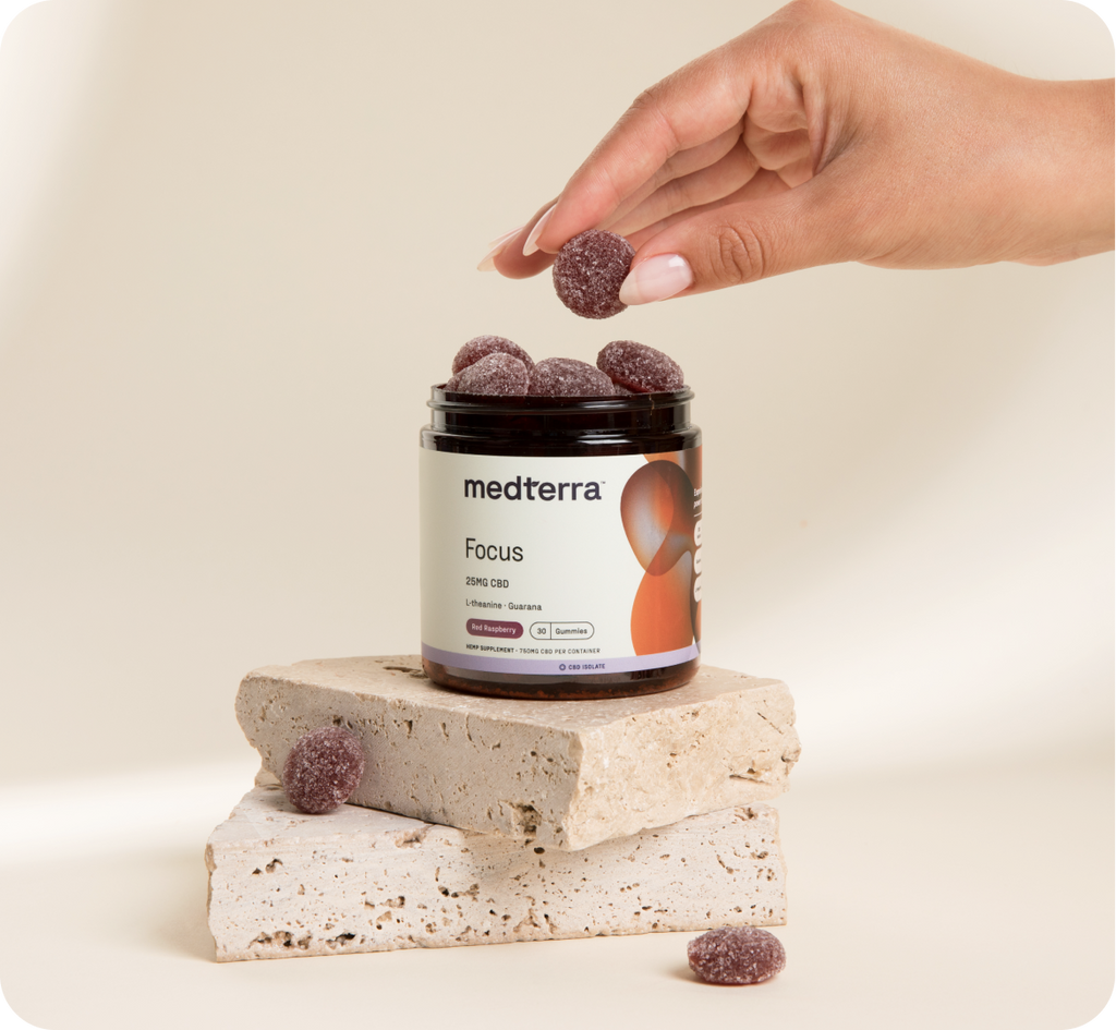 A hand is picking a Focus gummy from an open jar labeled "Medterra Focus 25MG CBD." The jar is placed on two stacked pieces of beige stone, with a few gummies scattered around. The background is a plain, soft cream color.