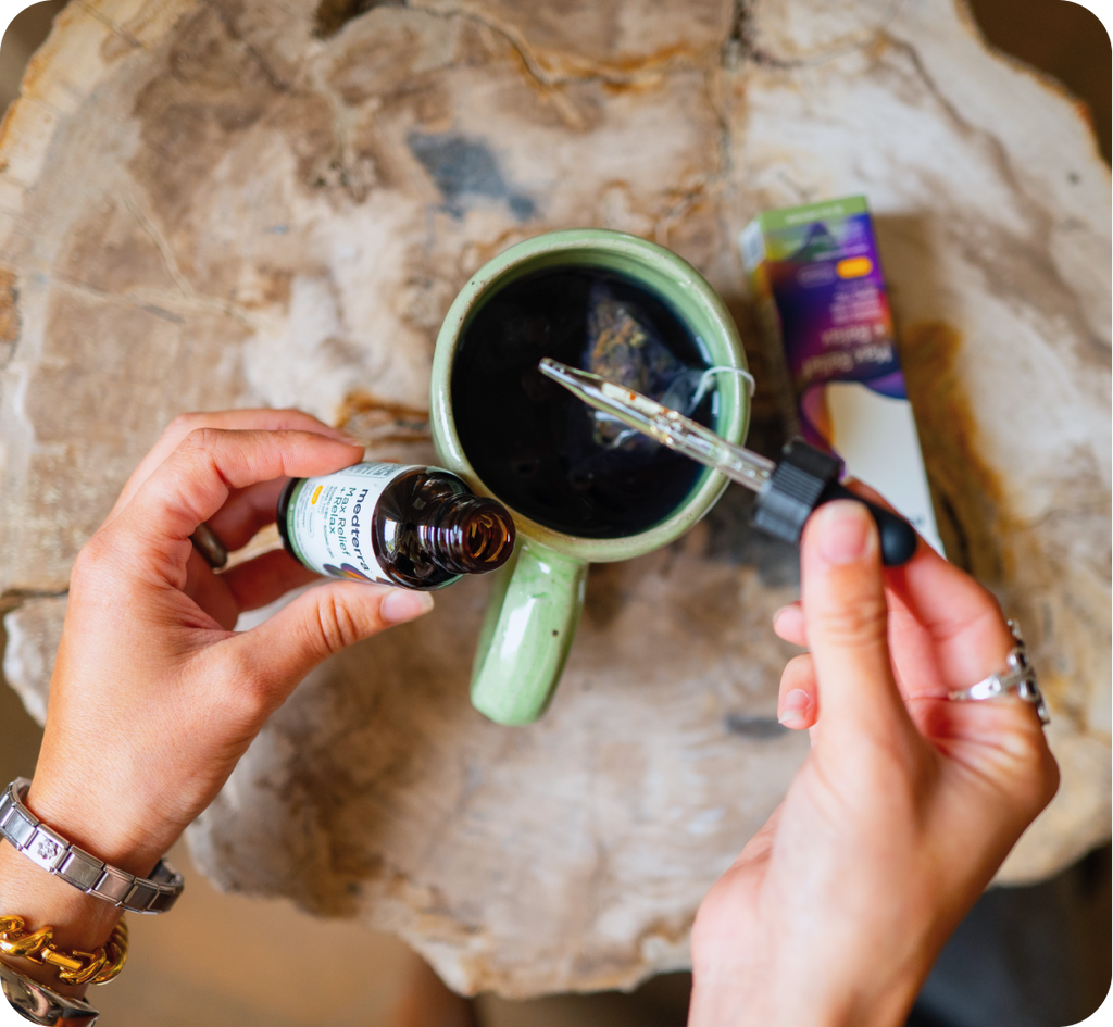 A person with bracelets on their wrist is holding a small tincture bottle with a dropper over a ceramic mug filled with a dark liquid. Nearby is a box labeled "MycoBotanicals" and "Immune Defense" with a purple accent. The scene is on a rustic wooden surface.