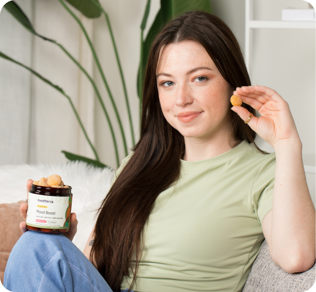 A woman with long dark hair, wearing a light green shirt, sits on a couch holding a round snack in one hand and a jar labeled "Mood Boost Gummies" in the other. There is a large plant in the background.