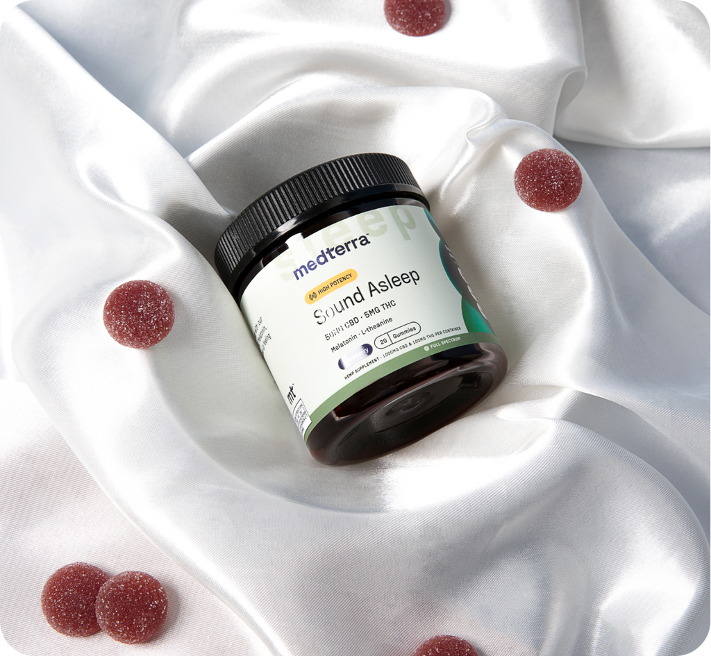 A jar of Medterra brand CBD+gummies, labeled "Stay Asleep," sits on a silky white fabric. Several red CBD sleep gummies are scattered around the jar, complementing the luxurious background. The jar and gummies are positioned to emphasize the product's relaxing attributes.