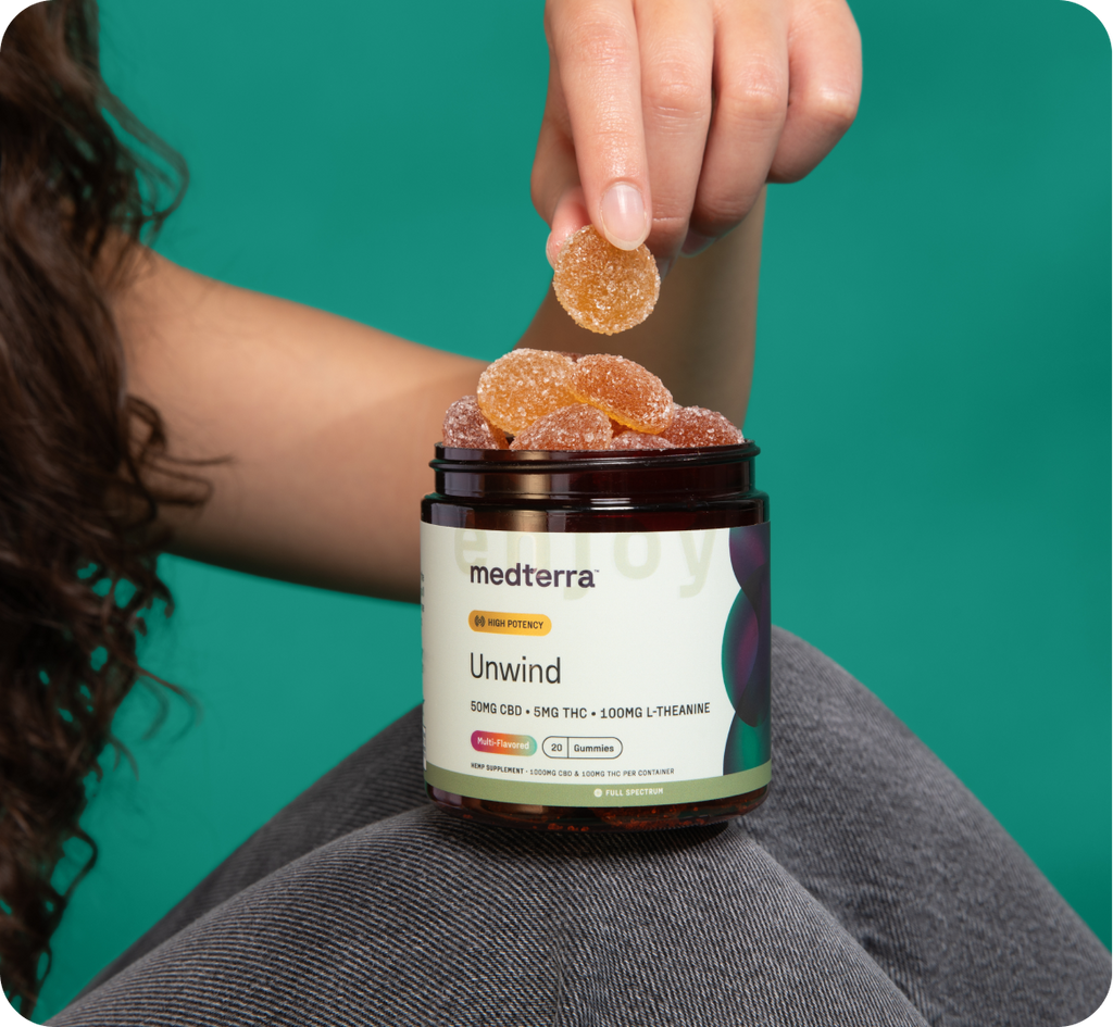 A person with long curly hair holds an open jar of Medterra Unwind Gummies, labeled with ingredients including CBD, THC, and L-Theanine. Against a teal background symbolizing tranquility, the person takes a gummy from the jar to experience deep calm.