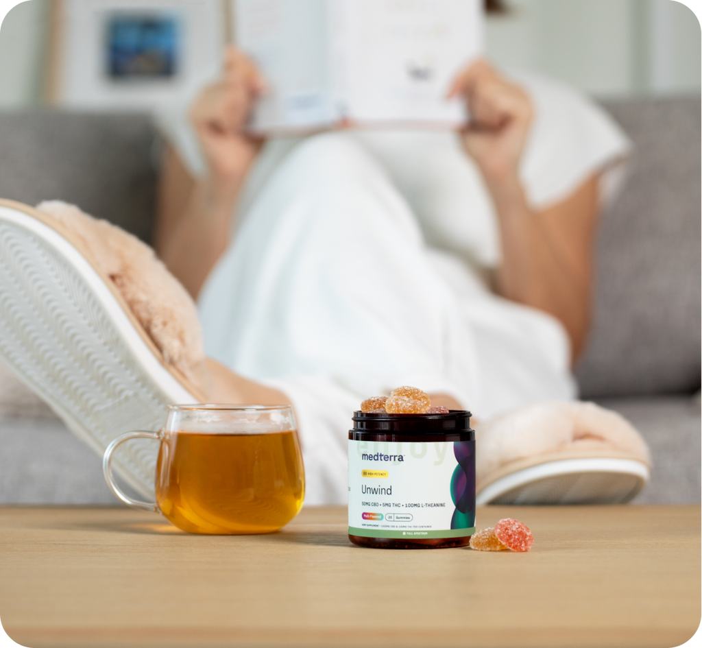 A person in cozy slippers relaxes on a couch, holding a book. In the foreground, there's a clear mug of tea and an open jar labeled "Medterra Unwind" with some tranquility-inducing gummies partly out on a table. The background includes blurred elements of a living room.